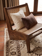 A mid-century modern wooden armchair with a woven cane back and seat, styled with two throw pillows (one cream, one dark brown) on a patterned area rug in a sunlit room.