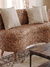 A curved woven sofa upholstered in a natural brown jute or seagrass material, styled with three light beige throw pillows in a warm, sunlit living room setting.