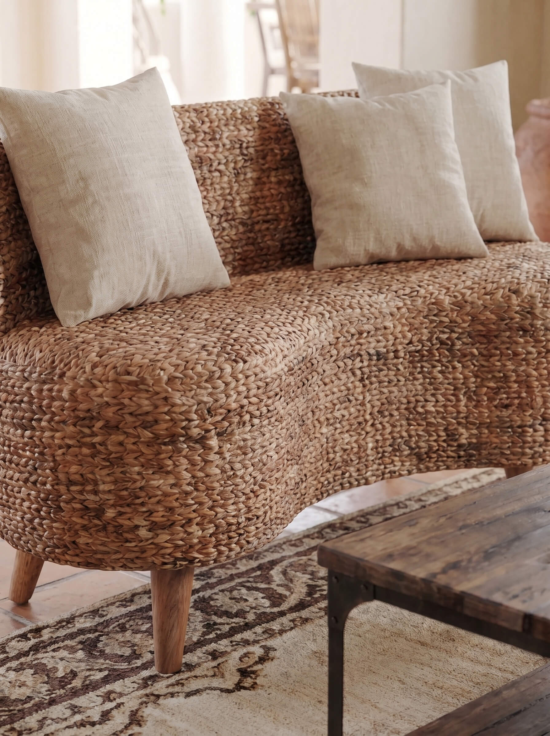 A curved woven sofa upholstered in a natural brown jute or seagrass material, styled with three light beige throw pillows in a warm, sunlit living room setting.