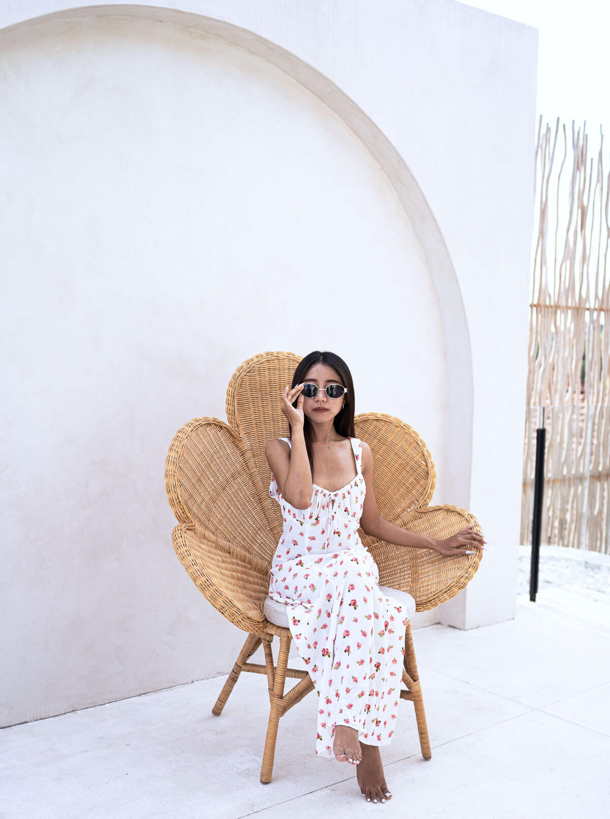 Woman in a white floral dress sitting on a wicker chair against a white wall.