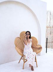 Woman in a white floral dress sitting on a wicker chair against a white wall.