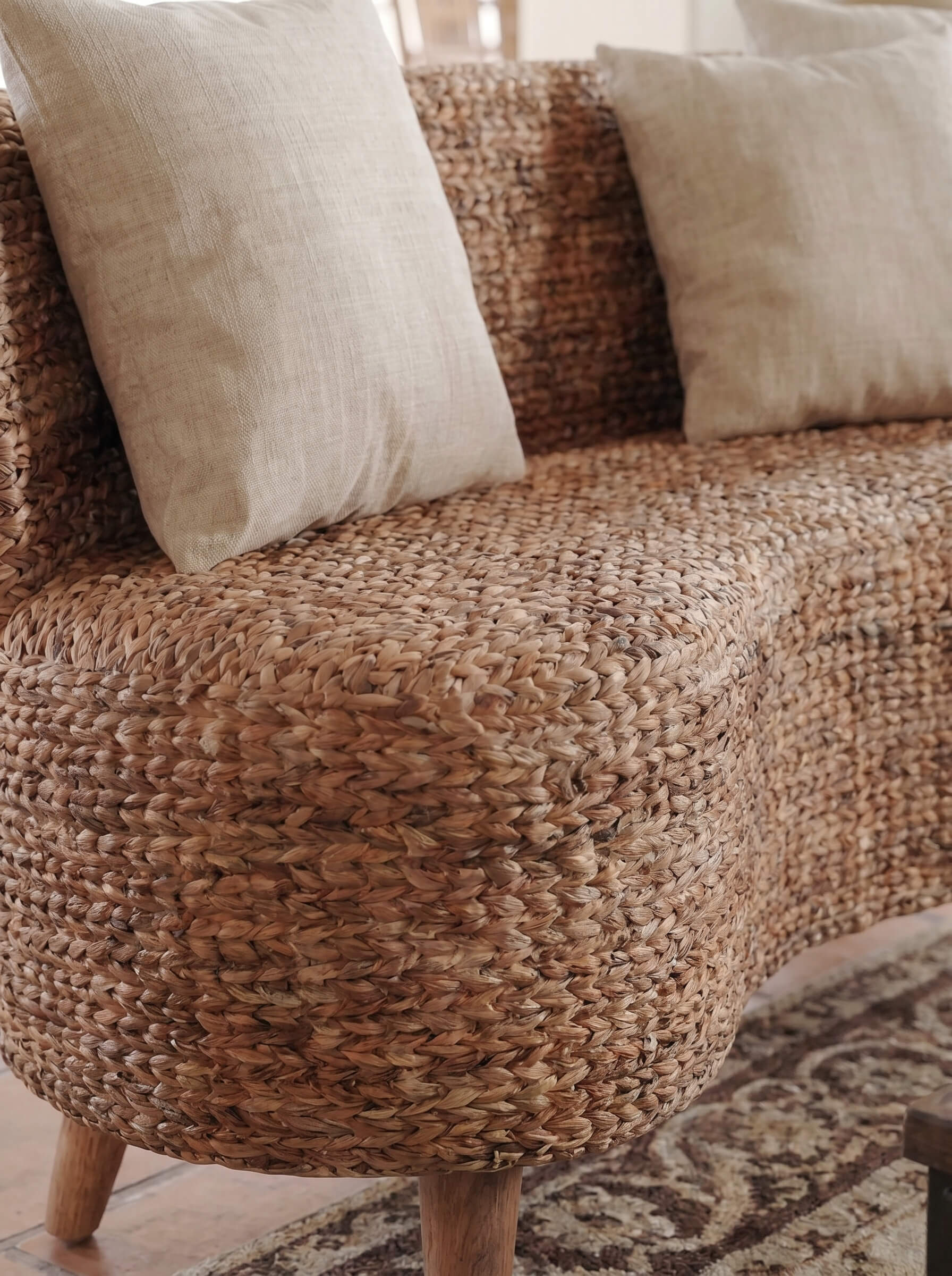 A close-up of a woven natural-fiber sofa or bench with two light beige throw pillows, shown in a softly lit indoor setting.