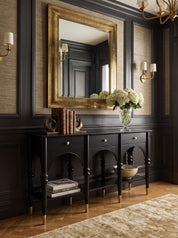 A black console table with gold legs and brass hardware, styled in a classic entryway under a large gold-framed mirror. The table holds a vase of white flowers and books, set against dark wood paneling and a light rug.
