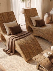 Two curved woven rattan chaise lounge chairs with cream and brown pillows and a brown throw blanket, styled in a warm, sunlit room with a rustic wooden table and neutral decor.