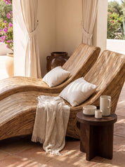Two curved woven rattan chaise lounge chairs with cream pillows and a beige throw blanket, styled on a terracotta tiled patio with a small wooden side table holding white ceramic mugs.