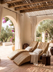Two curved wooden chaise lounge chairs with white pillows and a cream throw blanket, set on a tiled patio under a rustic wooden ceiling with arches opening to a garden.