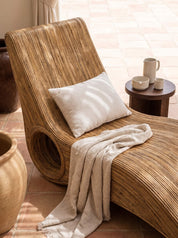 A curved woven rattan chaise lounge chair with a white throw pillow and a beige linen throw blanket, styled on a terracotta tiled patio next to a small wooden side table.