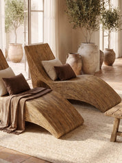Two curved woven rattan chaise lounge chairs in a warm, rustic indoor setting with potted olive trees, neutral throw pillows, and a brown throw blanket.