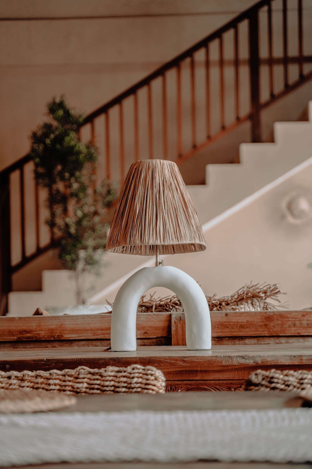 Decorative lamp with a textured shade on a wooden surface, with a staircase and plants in the background.