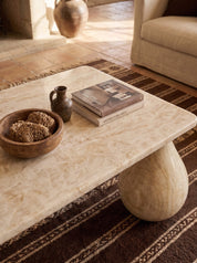 A rectangular coffee table made of light beige marble with a rounded sculptural base, styled in a warm living room with a brown patterned rug, a wooden bowl, and a stack of books.