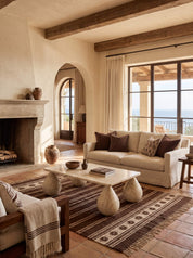A styled interior living room scene with a cream slipcovered sofa, patterned brown and cream pillows, a white stone coffee table with sculptural legs, a brown patterned area rug, terracotta tile flooring, a fireplace, exposed wood ceiling beams, and large windows overlooking water.