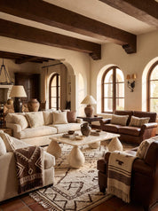 A styled living room interior with cream and brown sofas, a stone coffee table, patterned rugs, arched windows, and exposed wooden ceiling beams.