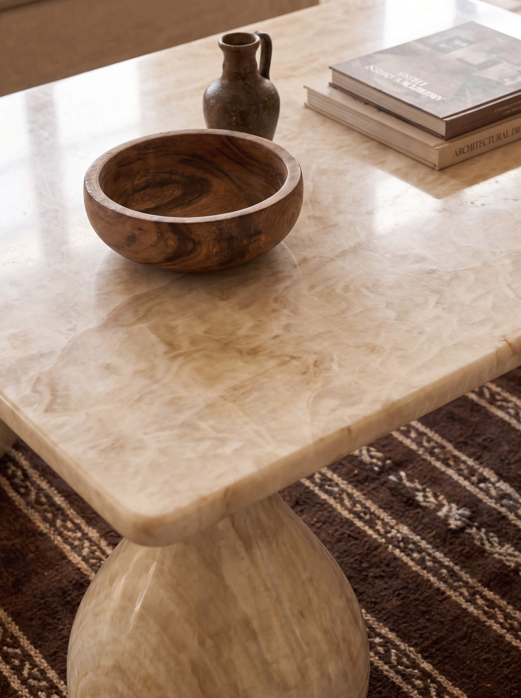A close-up of a polished beige marble or travertine table with a rounded bowl and stacked books on top, placed over a patterned brown rug.