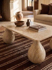 A rectangular coffee table made of polished cream-colored stone with rounded teardrop-shaped legs, styled in a warm, rustic living room with a brown patterned rug, a sofa, and decorative ceramic vases.