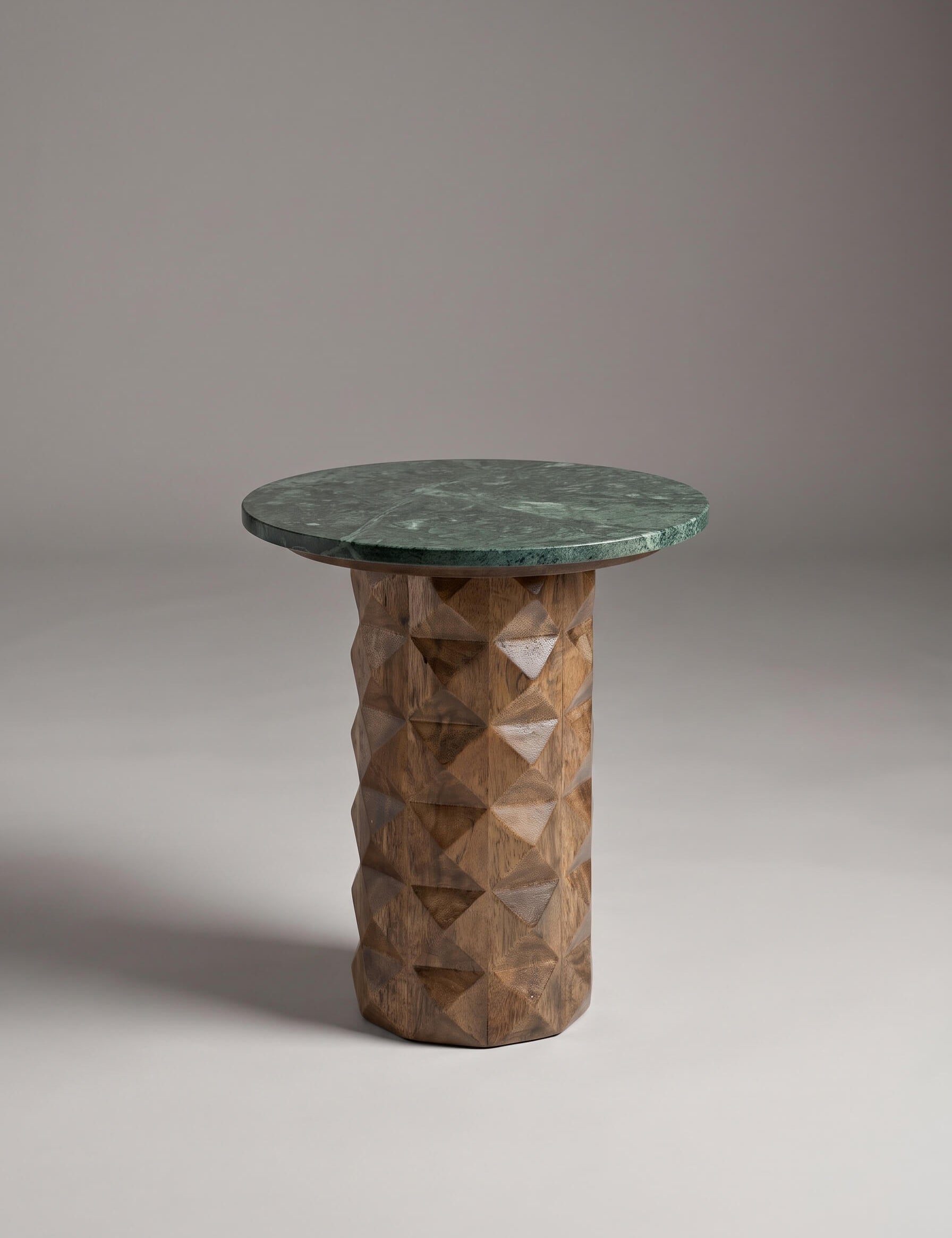 A modern side table featuring a round dark green marble top and a textured, faceted geometric wood pedestal base in a warm brown tone, set against a neutral gray studio background.
