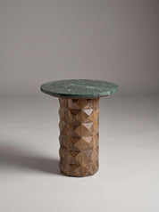 A modern side table featuring a round dark green marble top and a textured, faceted geometric wood pedestal base in a warm brown tone, set against a neutral gray studio background.