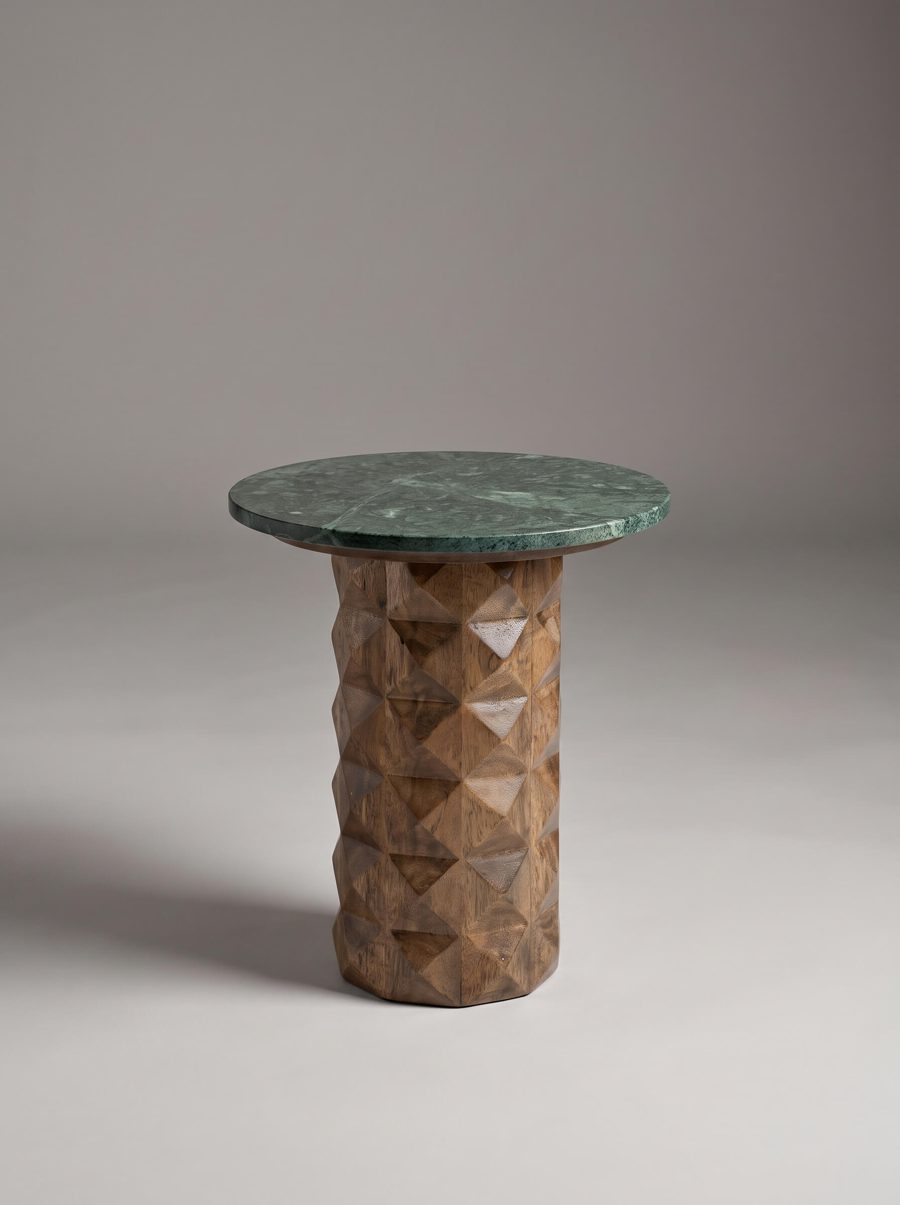 A modern side table featuring a round dark green marble top and a textured, faceted geometric wood pedestal base in a warm brown tone, set against a neutral gray studio background.