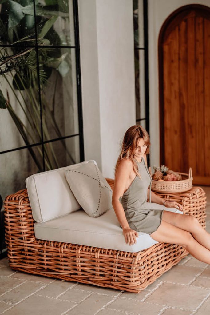 Woman sitting on a wicker chair with a baby in a basket, surrounded by plants and a wooden door.