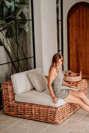 Woman sitting on a wicker chair with a baby in a basket, surrounded by plants and a wooden door.