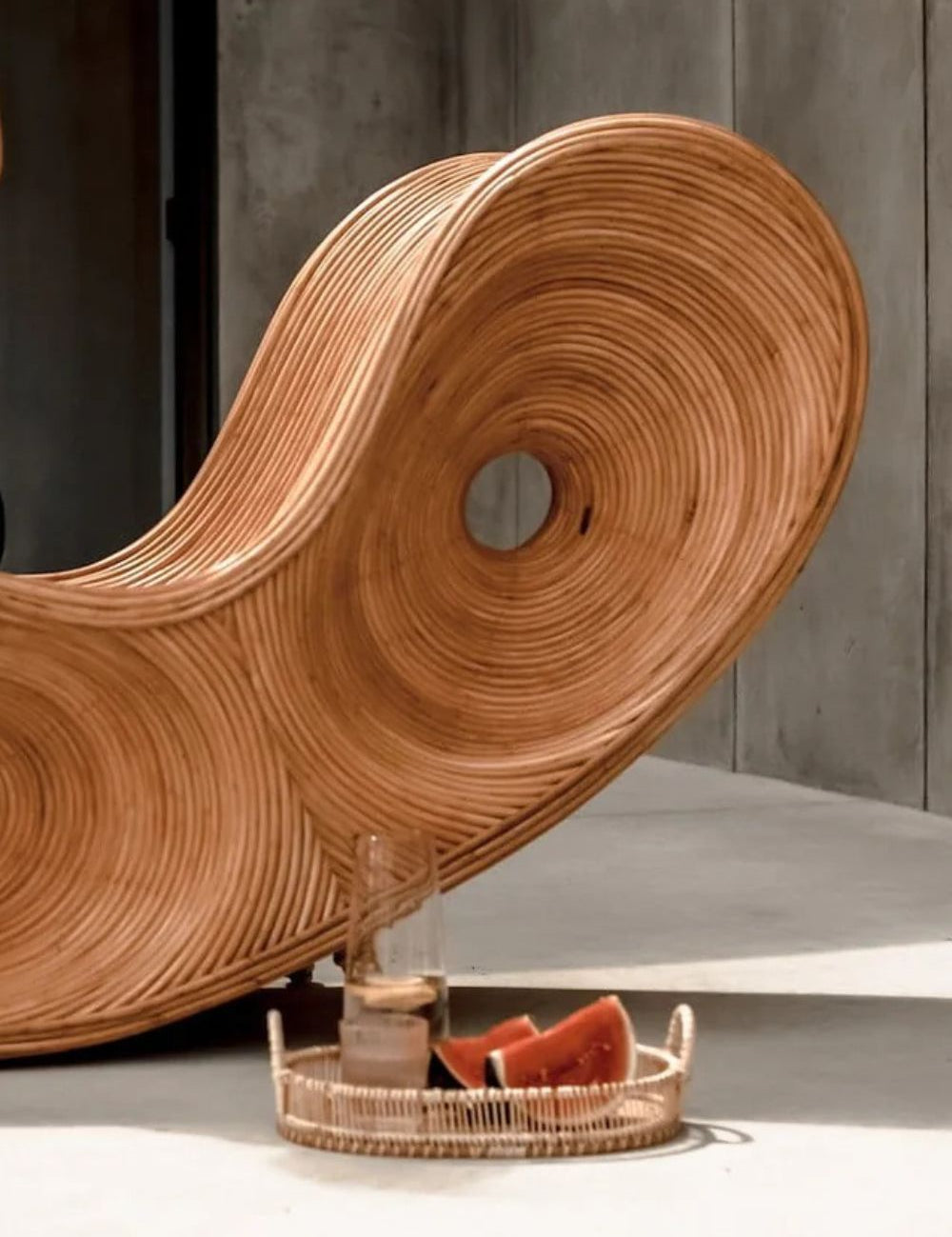Wooden wavy chair with a glass and fruit on a tray against a concrete wall.