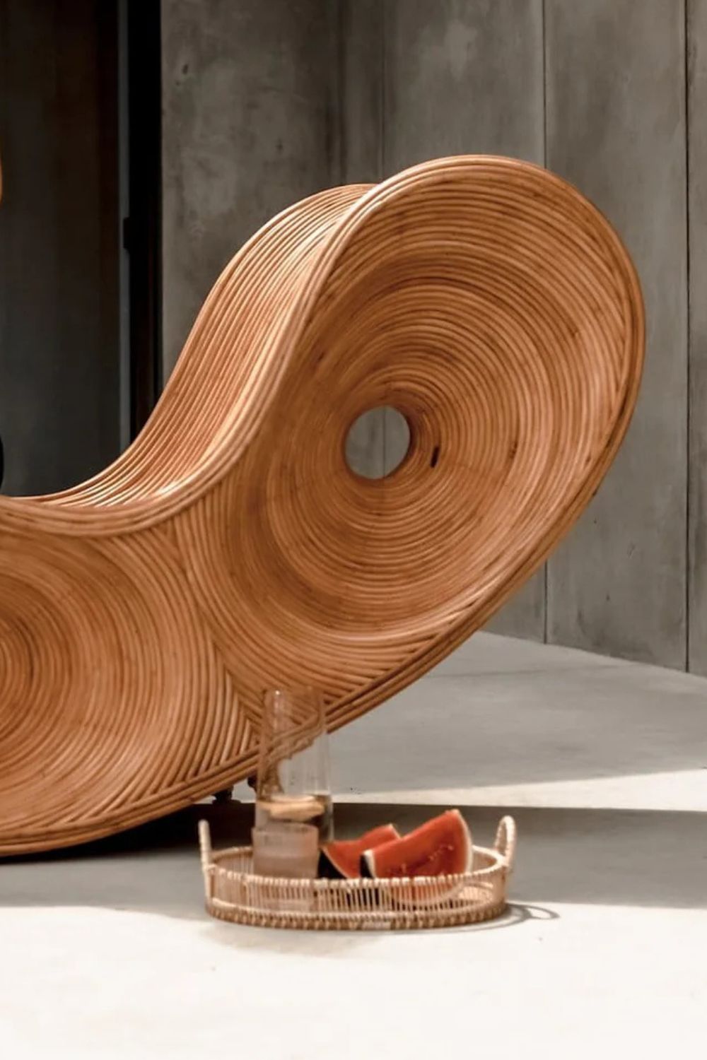 Wooden wavy chair with a glass and fruit on a tray against a concrete wall.