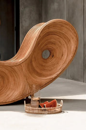 Wooden wavy chair with a glass and fruit on a tray against a concrete wall.