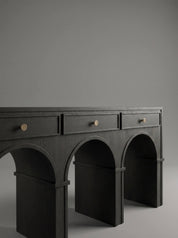 A dark charcoal gray console table with three drawers and three arched legs, featuring small round brass knobs, set against a solid gray studio background.