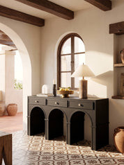 A dark charcoal gray console table with three arched legs and three drawers, styled in a rustic Mediterranean interior with a patterned tile floor, arched window, and wooden ceiling beams.