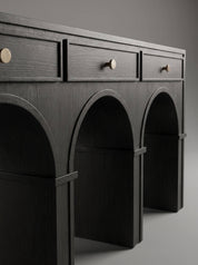 A dark charcoal gray wooden console table featuring three drawers on top with round brass knobs and three arched open shelves below, photographed against a neutral gray background.