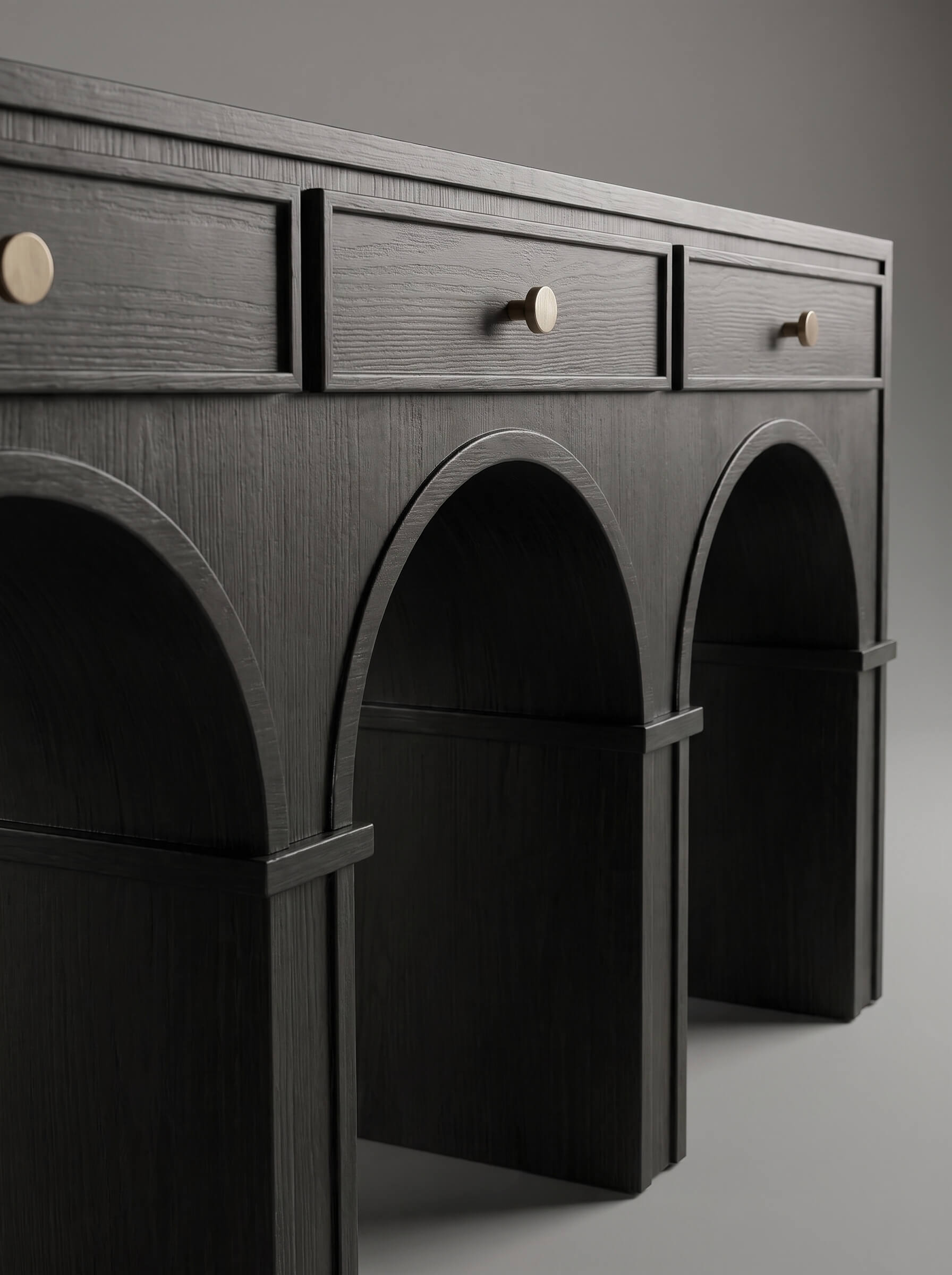 A dark charcoal gray wooden console table featuring three drawers on top with round brass knobs and three arched open shelves below, photographed against a neutral gray background.