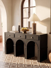 A dark charcoal gray console table with three drawers and three arched legs, styled in a warm, sunlit room with a patterned tile floor, a table lamp, and decorative items on top.