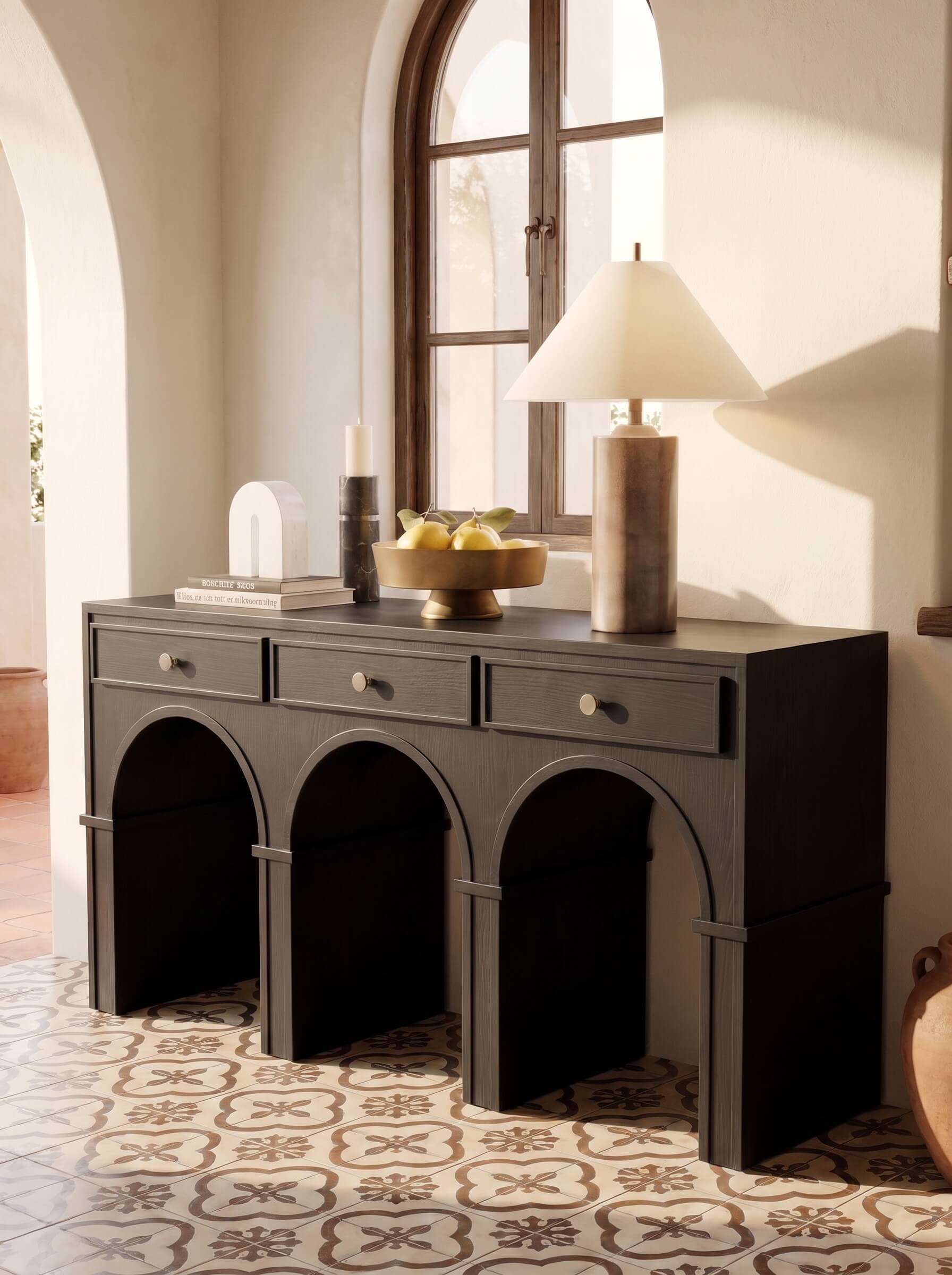 A dark charcoal gray console table with three drawers and three arched legs, styled in a warm, sunlit room with a patterned tile floor, a table lamp, and decorative items on top.