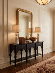 A black console table with gold-tone legs and drawer knobs, styled with two table lamps, books, and a bowl in a classic room setting with a large gold-framed mirror and patterned rug.