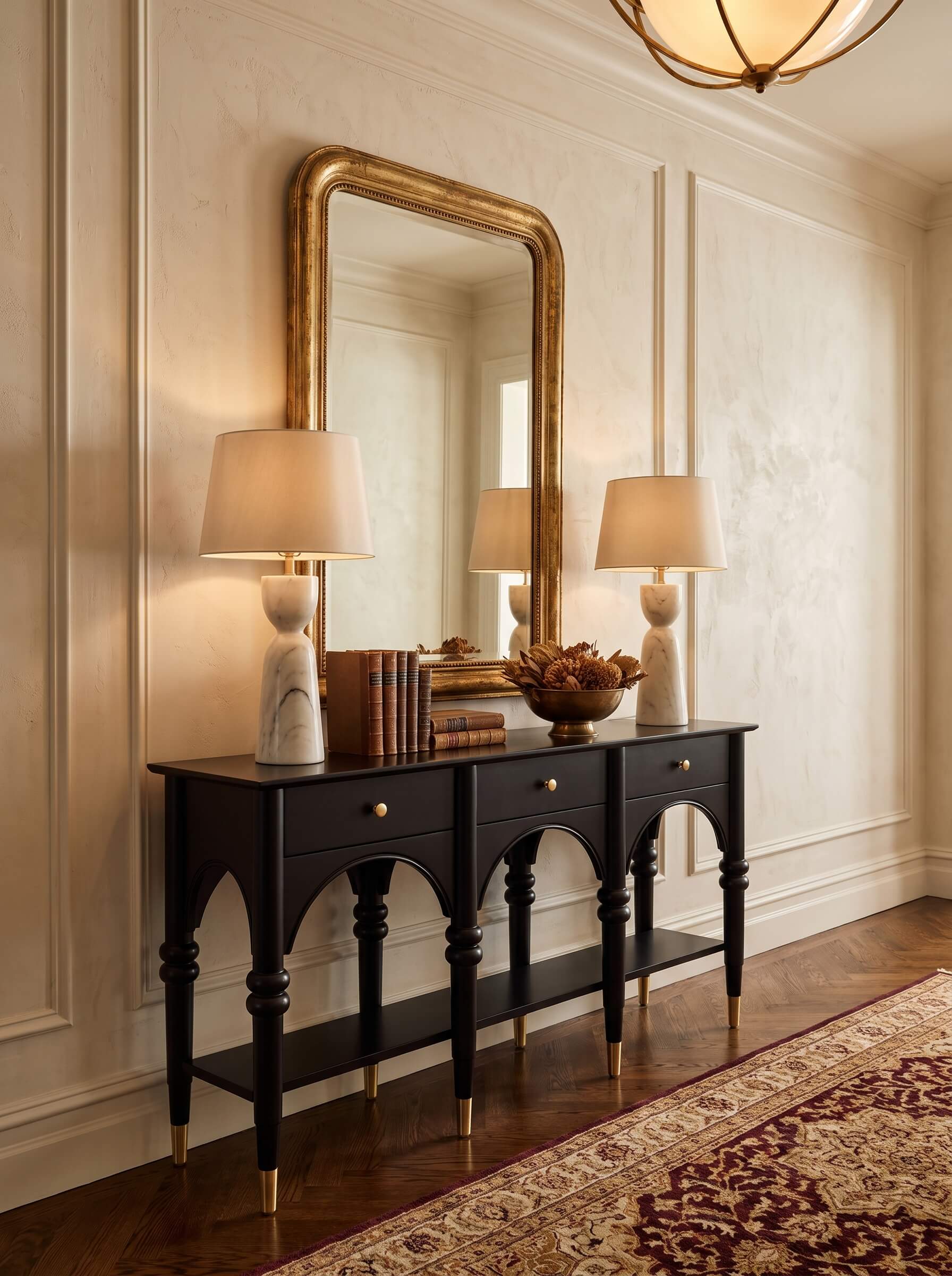 A black console table with gold-tone legs and drawer knobs, styled with two table lamps, books, and a bowl in a classic room setting with a large gold-framed mirror and patterned rug.