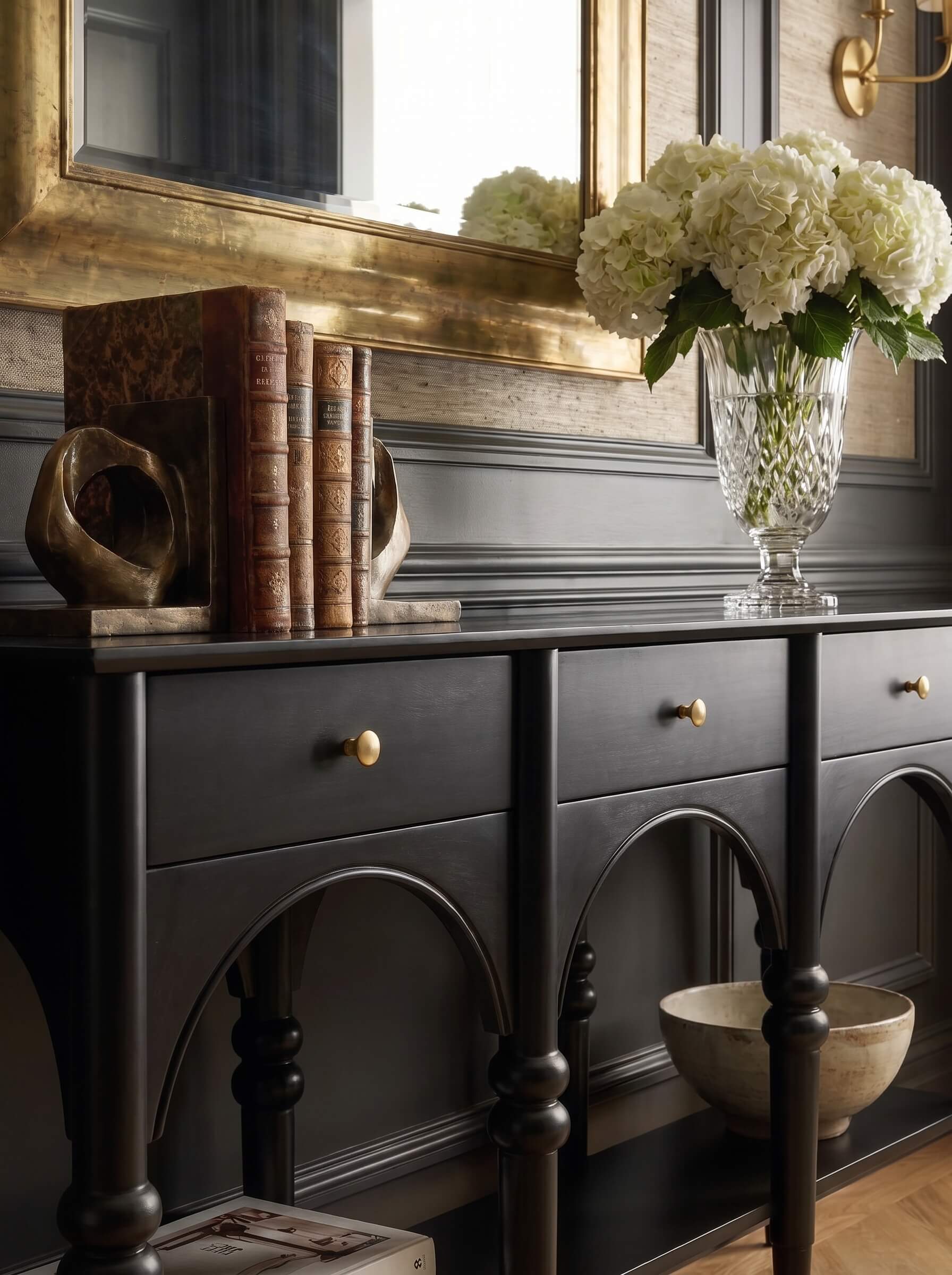 A dark wood console table with gold-tone drawer pulls and arched legs, styled in a room setting with books, a gold mirror, and a vase of white flowers.