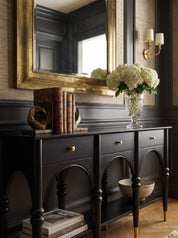 A dark wood console table with gold-tone hardware and legs, styled in an elegant room with a large gold-framed mirror, books, a vase of white flowers, and a bowl on a herringbone wood floor.