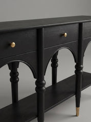 A close-up of a black wooden console table featuring arched drawer fronts, turned legs, brass drawer knobs, and a matching shelf, set against a neutral gray background.