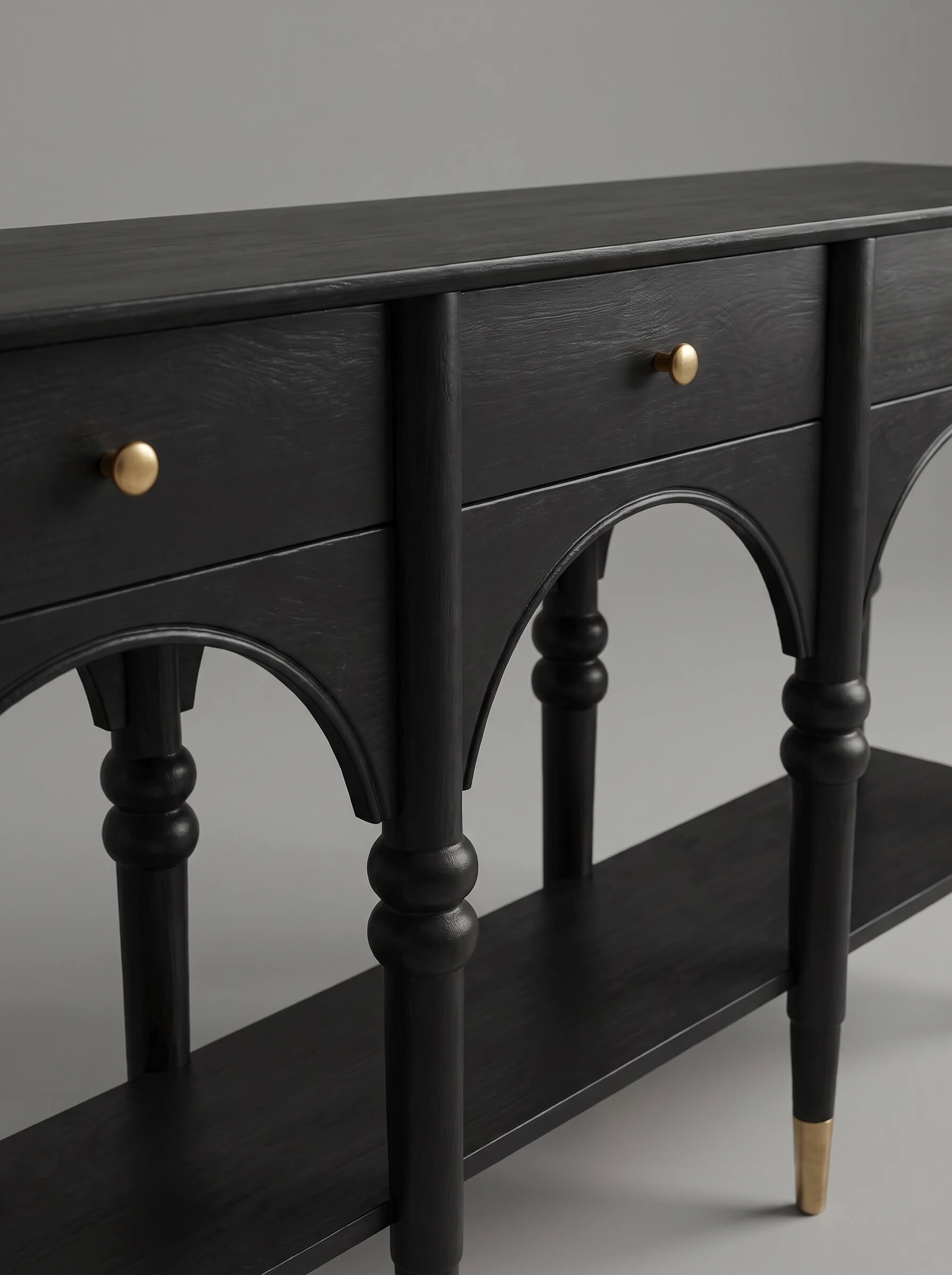 A close-up of a black wooden console table featuring arched drawer fronts, turned legs, brass drawer knobs, and a matching shelf, set against a neutral gray background.
