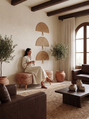A woman in a cream dress sits on a woven ottoman in a warm, rustic living room with dark wood ceiling beams, potted olive trees, and brown leather furniture.