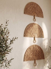Three natural woven rattan wall fans arranged vertically on a textured off-white wall, with soft sunlight casting leafy shadows and green plant leaves framing the scene.