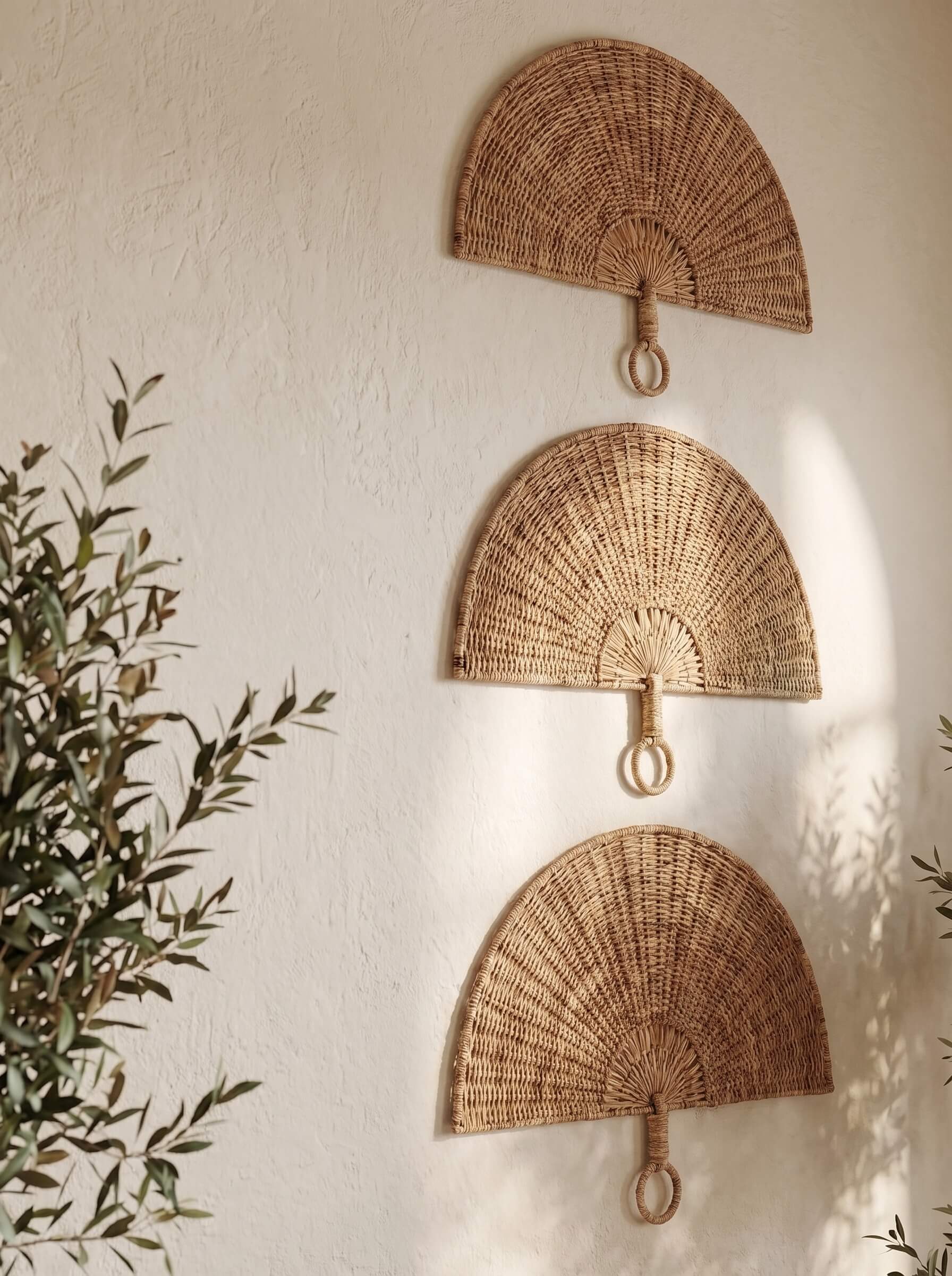 Three natural woven rattan wall fans arranged vertically on a textured off-white wall, with soft sunlight casting leafy shadows and green plant leaves framing the scene.
