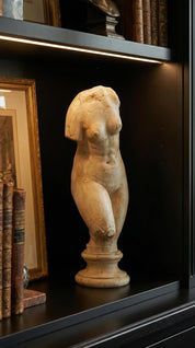 Stone sculpture of a nude figure on a dark shelf with books and a framed picture.