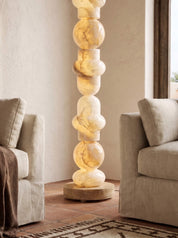 A tall sculptural floor lamp made of stacked warm white and cream marble spheres on a round wooden base, standing between two upholstered armchairs in a softly lit living room.