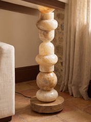 A sculptural floor lamp made of stacked polished stone spheres in warm cream and tan tones, mounted on a round wooden base in a styled interior room with curtains, tiled floor, and part of a sofa visible.