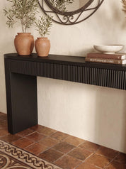 A modern black console table with a fluted front and open pedestal leg, styled in a warm interior with terracotta vases, books, and a bowl against a textured cream wall and tiled floor.