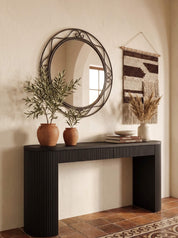A black ribbed console table in a warm, bohemian-style room, styled with two terracotta planters, a round black metal mirror, a woven wall hanging, books, and a small vase.