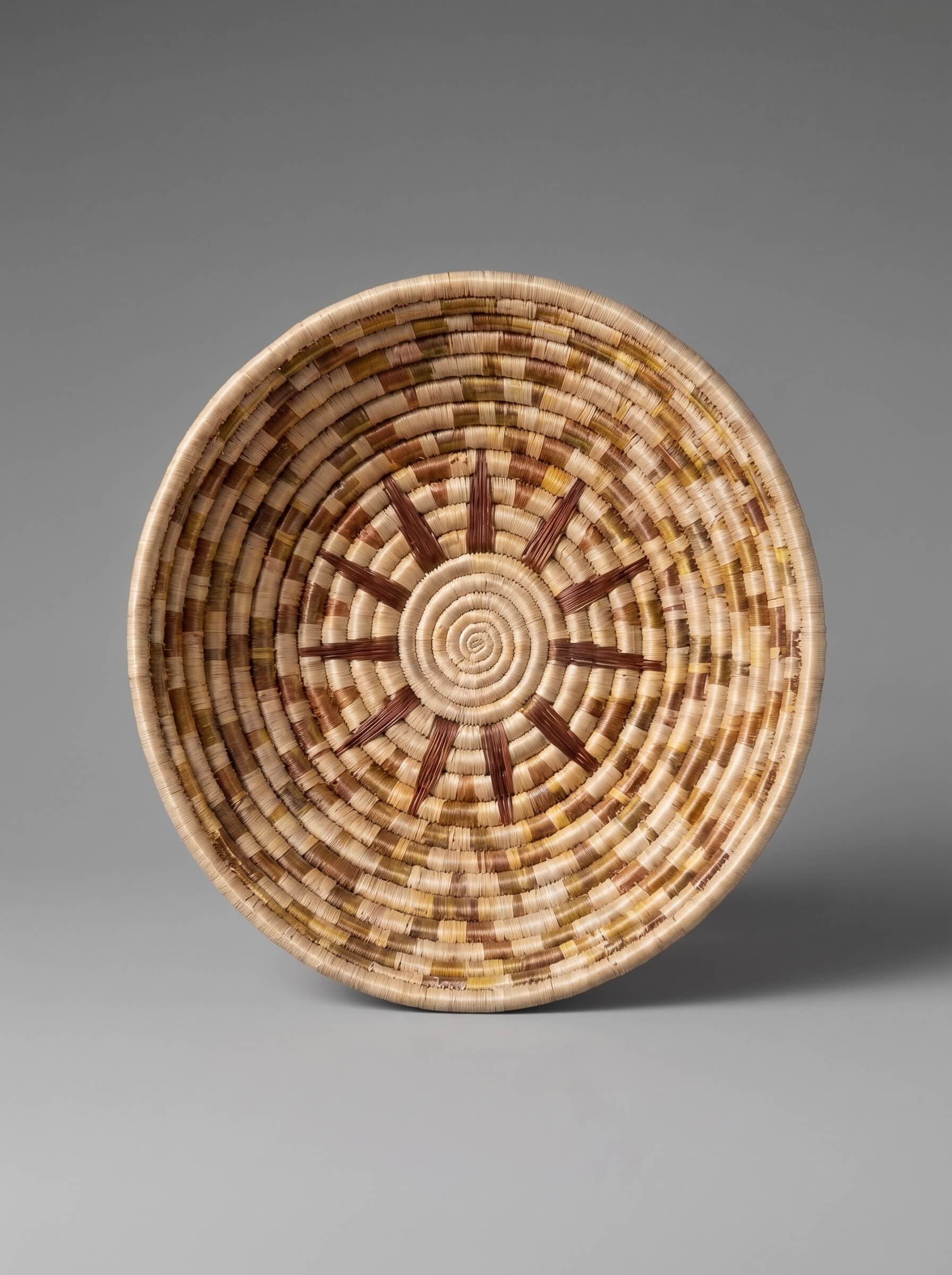 A hand-woven basket bowl with a geometric sunburst pattern in natural tan, beige, and brown tones, photographed against a neutral gray background.