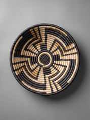 A round woven basket with a geometric spiral pattern in black and natural tan, photographed from directly above against a neutral gray background.