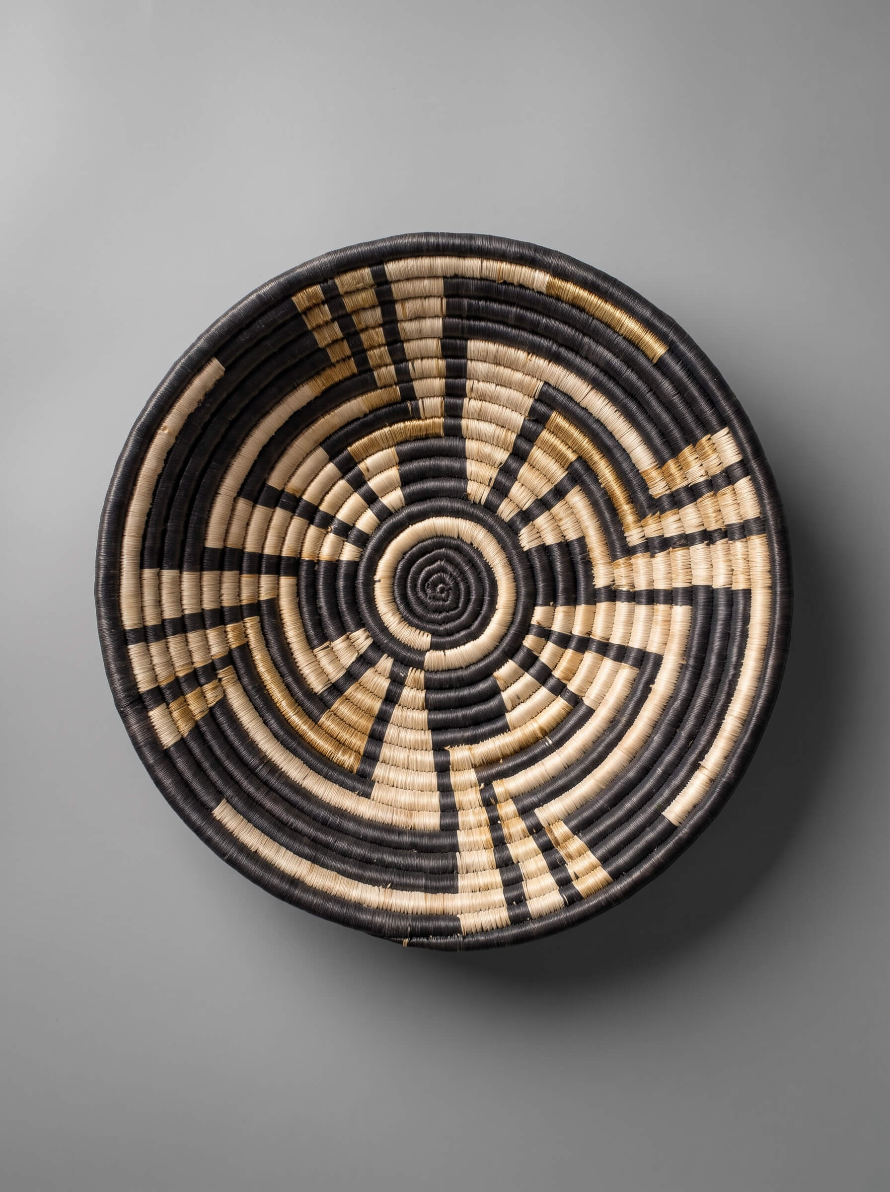A round woven basket with a geometric spiral pattern in black and natural tan, photographed from directly above against a neutral gray background.