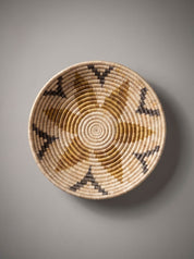 A round woven basket wall hanging with a geometric sunburst pattern in natural tan, brown, and black, shown against a neutral gray background.